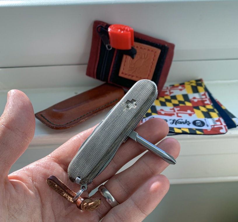 Shows the dedicated Philips head screwdriver on the Victorinox Tinker with olive drab green micarta scales in the foreground with EDC gear in the background